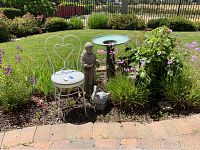 Garden scene with white iron chair with heart shape backrest, stone saint statue, small duck statue, and bird bath on pedestal surrounded by plants and flowers.