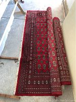 Full view of the partially unrolled red Bokhara rug showing overall pattern and fringe edges on a concrete floor.
