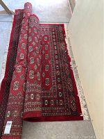 Hand tied Bokhara rug partially rolled out showing red patterned design and fringe along edges.