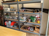 Photo displaying shelving unit with multiple plastic bins containing assorted garden tools and accessories, hand tools, plant pots, decorative flags, and wire stakes.
