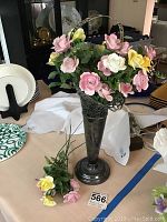 Full view of the tall metal vase holding porcelain flowers in pastel colors on a table with other dishes and knick-knacks.