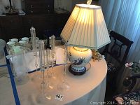 Photo of Asian ceramic lamp with cream color pleated shade and five clear glass oil candlesticks on white table.
