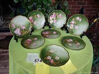 Eight scalloped porcelain plates displayed on a green tablecloth. Plates have green gradient background with pink floral designs and gold trim.