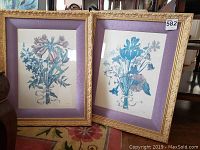 Pair of botanical prints in gold wooden frames with purple silk matting and glass fronts, framed and standing on floor.