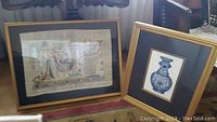 Two framed art prints on the floor near a window. One is a signed print with figures, the other is an Egyptian art of a blue ceramic vase.