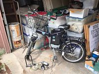 Front side angled view of black women's electric bike with metal basket on handlebars, charger and cable lock on floor nearby, stored in a cluttered garage.