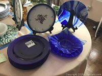 Photo showing 8 blue glass 10 inch dinner plates stacked, a bone china plate with blue grape design on stand, and two decorative blue and transparent glass plates on stands.