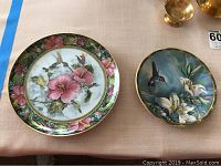 Two collectors plates on a table: the Pickard plate with hummingbird and pink flowers, and the Franklin Mint plate with hummingbird and white lilies.
