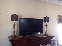 Photo showing the Curtis brand 36 inch flat-screen television centered on a wooden cabinet flanked by two ornate table lamps with dark brown shades.