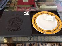 Black box with embossed Versace Medusa logo and branding on the top, showing the plate inside on a table with books.