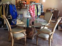 Wide view of round glass dining table with four upholstered wood chairs around it, set indoors among other furniture and clothing racks.