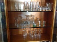 Various barware glasses displayed on two glass shelves inside a wooden cabinet.