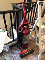 Red and black upright Bissell vacuum cleaner with cord wrapped around handle, placed near stair railing indoors
