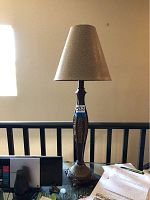 Full view of one of the two decorative table lamps, showing carved dark wood base with intricate detailing and beige tapered lampshade, approx 34 inches tall, placed on black railing.