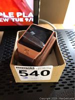 Smartwatch with a square black screen, rose gold casing, and brown strap resting on cushion inside box.