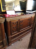 Front and side view of the dresser showing ornate gold trim, brown wood finish, black metal drawer handles, and minor scratches.