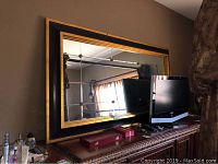 Frontal view of the large wall mirror resting on a dresser, showing the gold and black frame, and parts of a TV and other items nearby.
