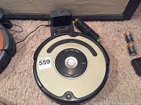 Top view of iRobot Roomba vacuum cleaner with charging dock, power cord, brush roller, and batteries beside it on carpeted floor.