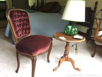 View of mahogany chair with burgundy velvet tufted seat and back with carved wooden frame, next to mahogany side table with green glass lamp on top.