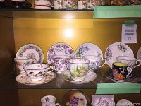 Front display of 7 china cups and saucers with various floral patterns and one black lacquerware demitasse set.