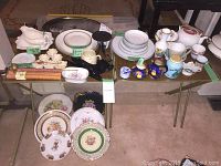 Photo of two tables displaying assorted china and household items including plates, cups, saucers, pitchers, and decorative pieces arranged in groups by brand and pattern.