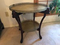 Side view of two tier round brass tray table with carved teak wood stand, showing front corner and decorative leg detail.