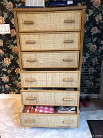 Front and side angled view of tall bamboo hutch with textured woven rattan drawers, some drawers open showing contents.