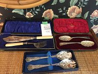 Full lot overview showing two boxes: one blue lined with cake server utensils (knife, fork, server) with ivory colored handles; the other red lined fitted case holding two ornate silver plated berry spoons with floral and scroll designs.