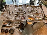 Overview of assorted service cutlery spread on a white cloth, including spoons, forks, knives, ladles, and several napkin holders.