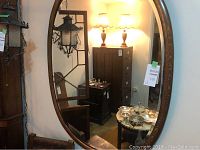 Full view of the large oval mirror hanging on the wall reflecting a room with furniture and lamps.