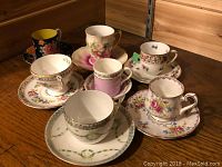 Seven assorted china teacups and saucers arranged on a wooden surface showing various patterns and colors.