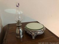 Overall view of the pewter lantern with glass chimney and tabletop mirror with metal feet on wooden table.