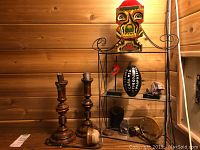 Photo showing overall lot with wooden candlesticks, metal and glass shelf, and various decorative objects including painted mask and stones.