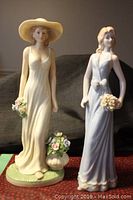 Front view of two porcelain female figurines, one with cream dress and hat, other with lavender dress