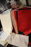 Assortment of folded cloth napkins, eight tablecloths in various colors and patterns including cream, red and green, and a red table runner with scalloped edges laid out on surface