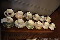 Twelve cup and saucer sets arranged on a wooden surface, showing various floral patterns and gold trim