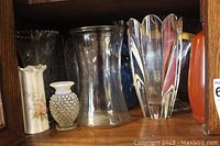Collection of assorted vases on a wooden shelf showing variety of clear glass, crystal, frosted, and colored vases.