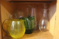Four glass pitchers and one glass decanter on wooden shelf. Yellow and green pitchers in front.