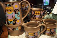 Full view of creamer, sugar bowl, and stacked cups showing owl and floral design on a brown ceramic base.