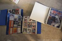 Three blue binders positioned on carpet, one binder open showing cards in plastic sleeves; the Beckett Tribute Nolan Ryan magazine visible at the front of one binder.