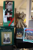 Photo of five Christmas decorations including Santa in box, angel figurine, Christmas caroler hurricane candle holder, and boxed decorative houses.