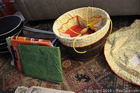 Three folding stools with black seats and metal legs, folded and stacked on the left. Half barrel picnic basket with yellow floral liner and yellow cord handles. Picnic plastic tablecloths and accessories laid out on a patterned carpet.