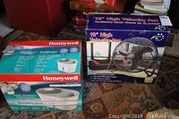 Lot items including boxed FC 12-inch High Velocity Fan and boxed Honeywell Cool Moisture Humidifier placed side by side on carpeted floor.