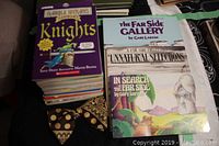 Stack of books including Horrible Histories title Knights, and several The Far Side books by Gary Larson, with a polka dot clip-on bow tie placed in front