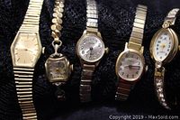 Five ladies goldtone wristwatches displayed on black fabric, varying case shapes and band styles.