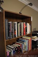Front and side view of wood-like bookshelf filled with books and attached black adjustable lamp.