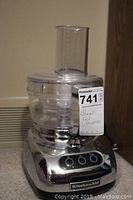 Front view of KitchenAid food processor showing clear plastic bowl, feeding chute, and chrome base with buttons