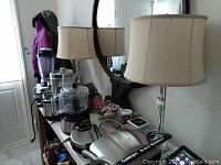 Pair of table lamps on a surface with kitchen appliances visible in the background. Both lamps have cream shades and clear glass and silver metal bases.