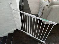 White metal expandable baby gate installed at the base of a stairway next to a white brick wall and living room area.