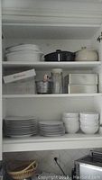 Photo shows shelves holding stacked white ceramic plates, bowls, casserole dishes in white, black, and cream colors, metal tins, glass canister, and metal decorative bin.